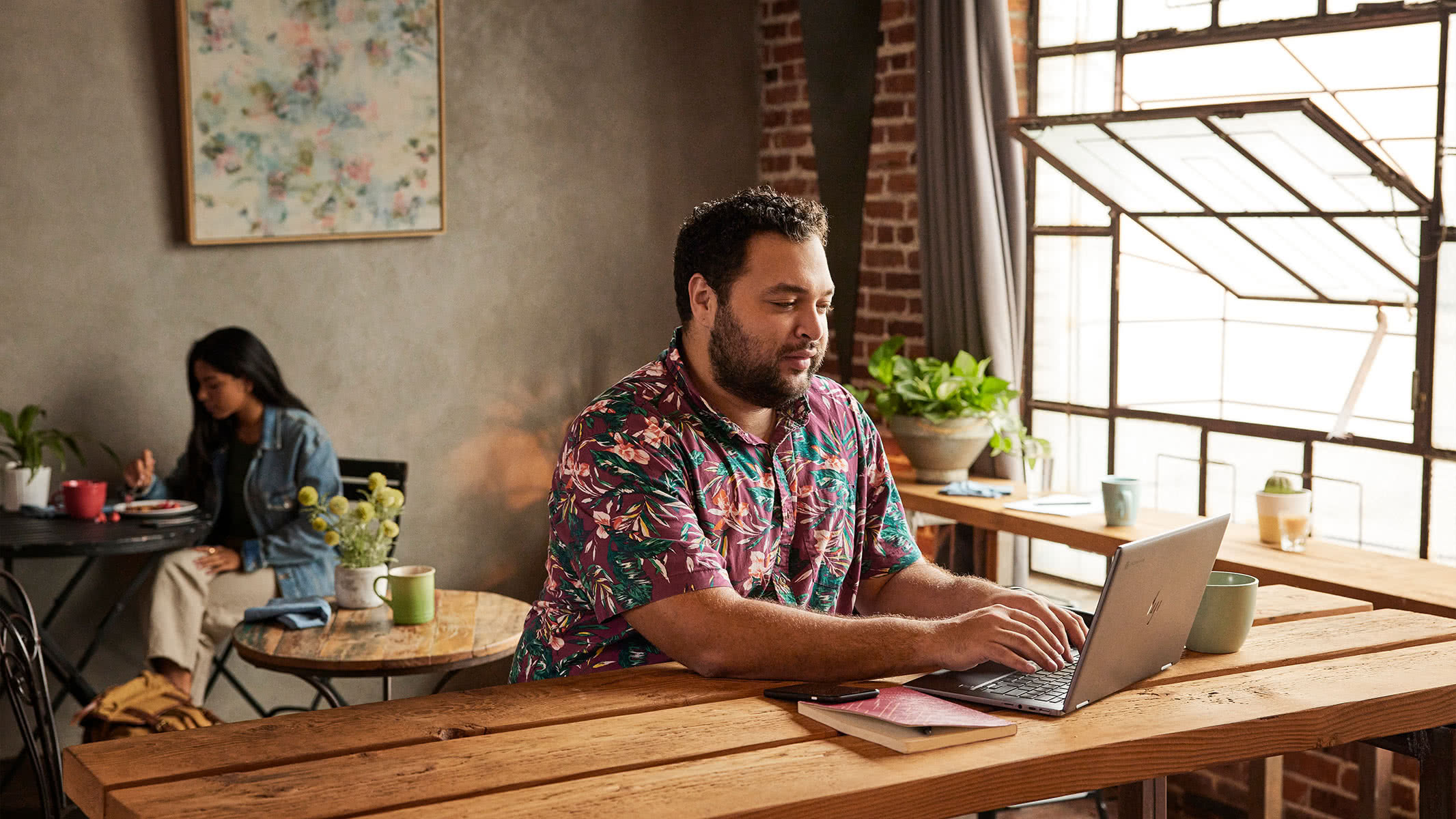 A person stands over a desk holding a mug in their right hand and touching an open Chromebook screen with their left hand.