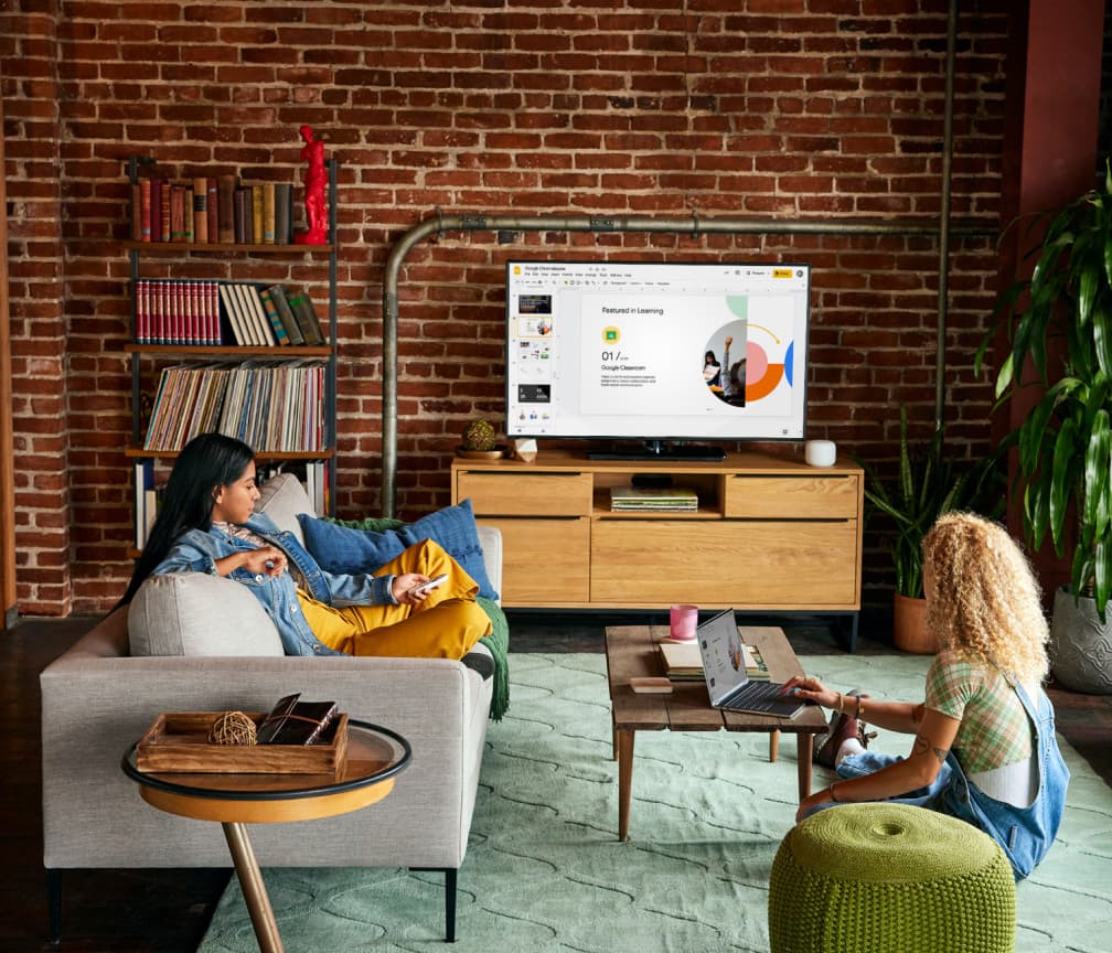 A person sits on a couch watching a TV presentation. A second person sits on the floor at a coffee table with a Chromebook.