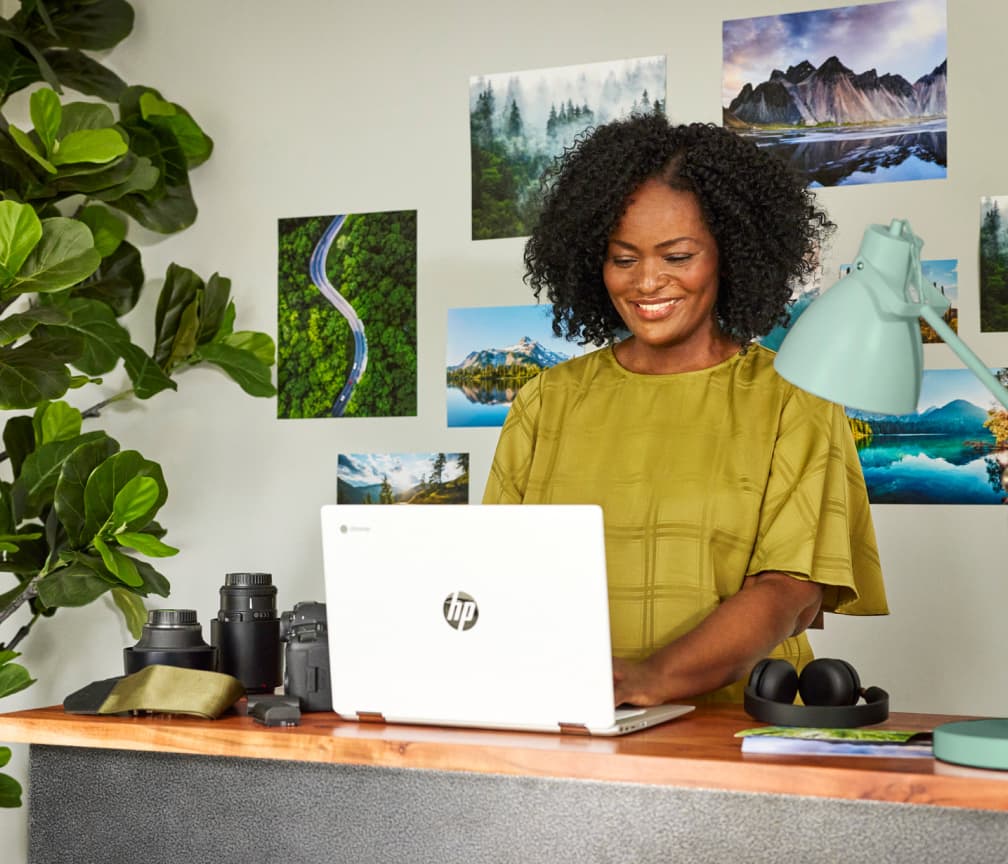 A person stands in front of a wall of photos and types on a Chromebook that sits on a table surrounded by camera lenses.